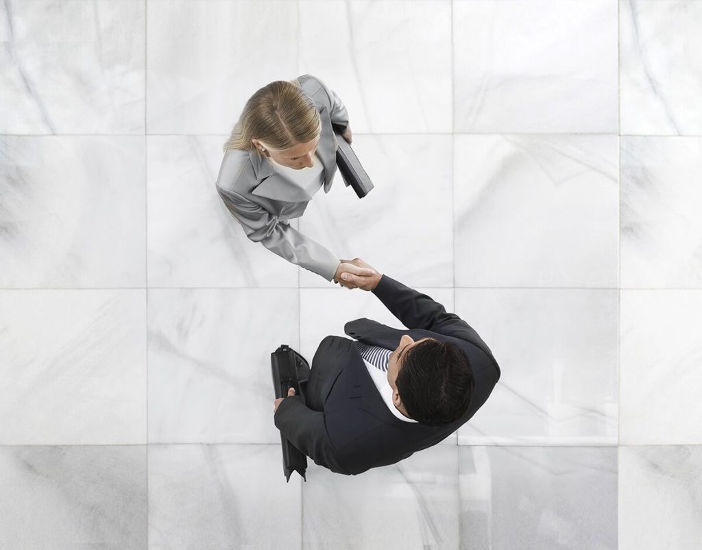 Overhead view of two businesspeople in formal attire shaking hands on a tiled floor, each holding a folder.