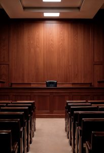 Empty courtroom with wooden walls, rows of seats facing a judge's bench, and a single black chair behind the bench.