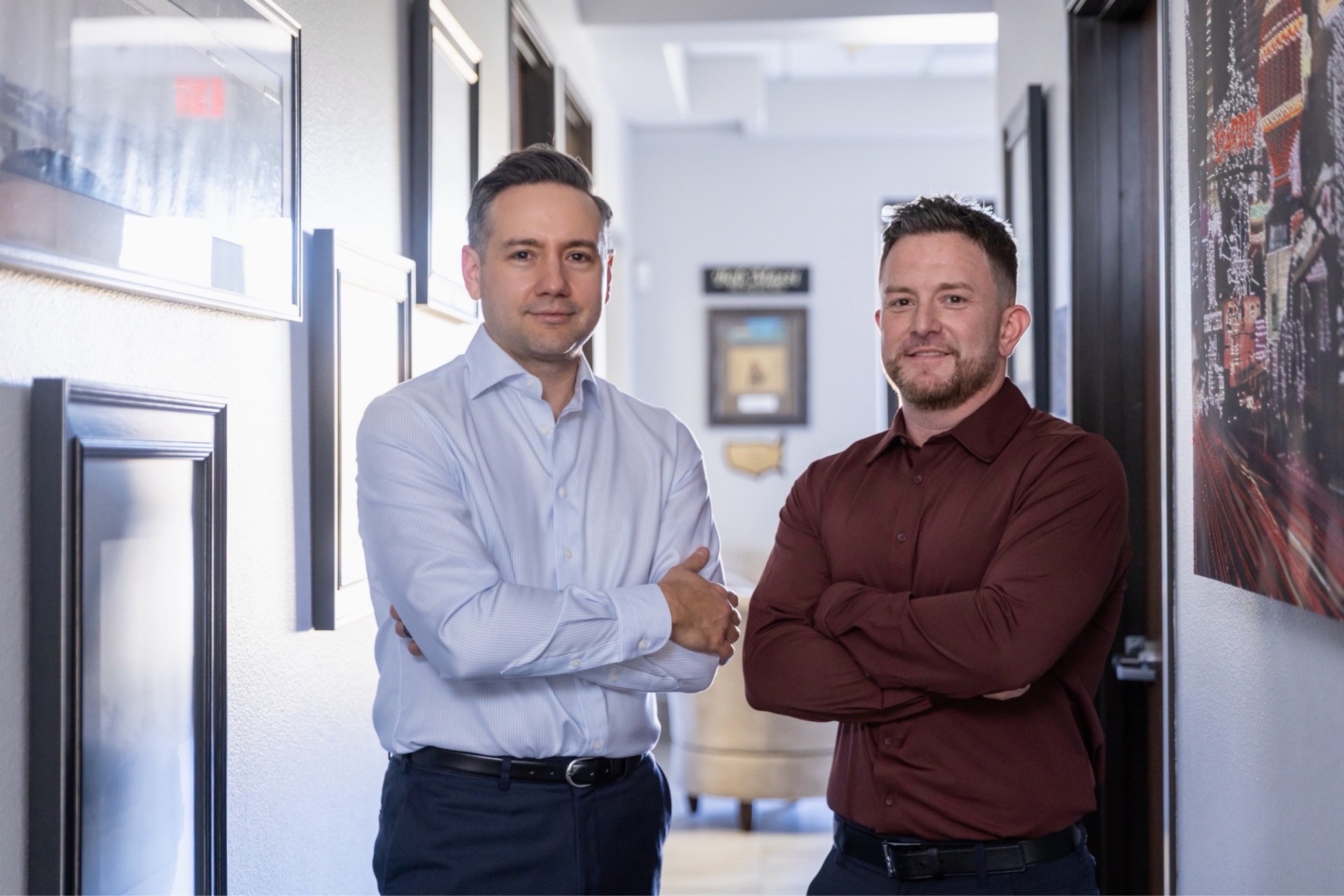 Two men stand side by side in a hallway with framed pictures on the walls, both with arms crossed and looking at the camera.